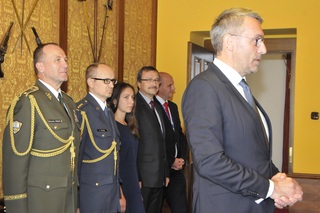 Ministr obrany při svém krátkém projevu, za ním čestní hosté: brigádní generál Bohuslav Přikryl, rektor-velitel Univerzity obrany a předseda vědecké rady Univerzity obrany, plk. gšt. Alexandr Štefek, prorektor pro vědeckou a expertní činnost, Nora Boštíková, dcera Vandy Boštíkové, a Miroslav Špliňo z Fakulty vojenského zdravotnictví Univerzity obrany